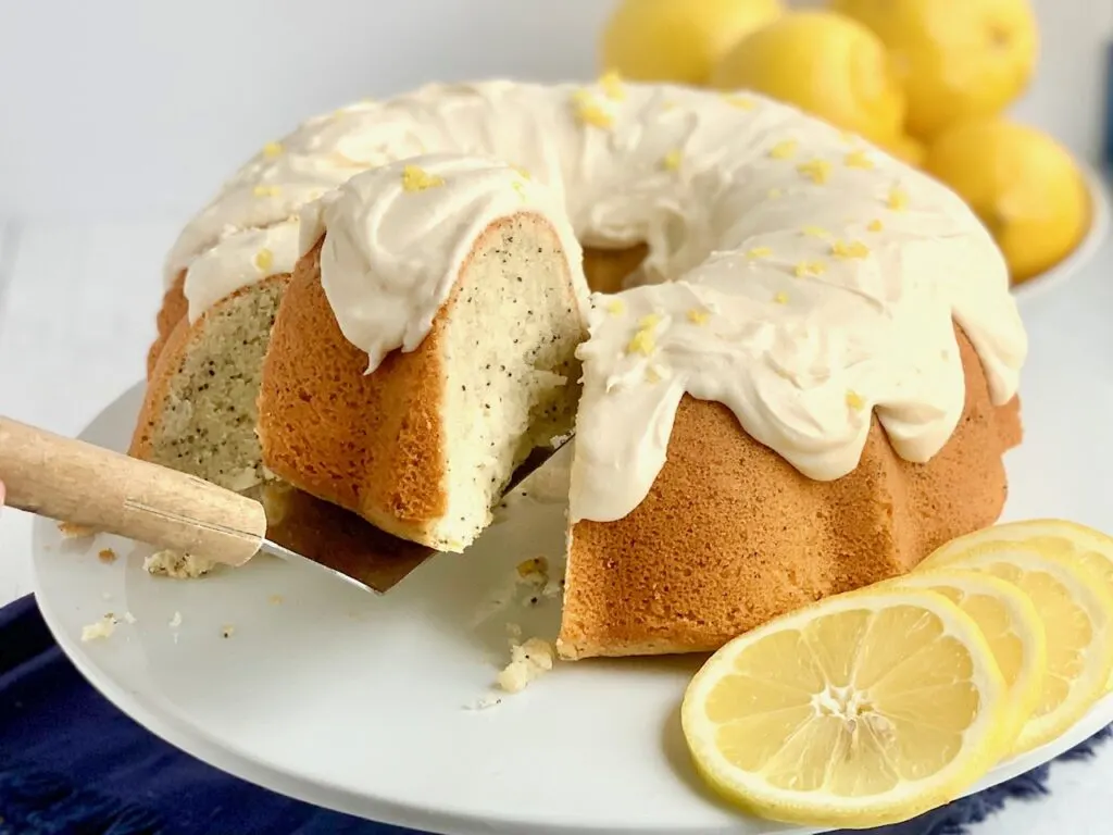 A round lemon poppy seed bundt cake iced with a thick lemon cream cheese spread ad topped with fresh lemon zest. a slice has been cut and is being lifted from the cake.