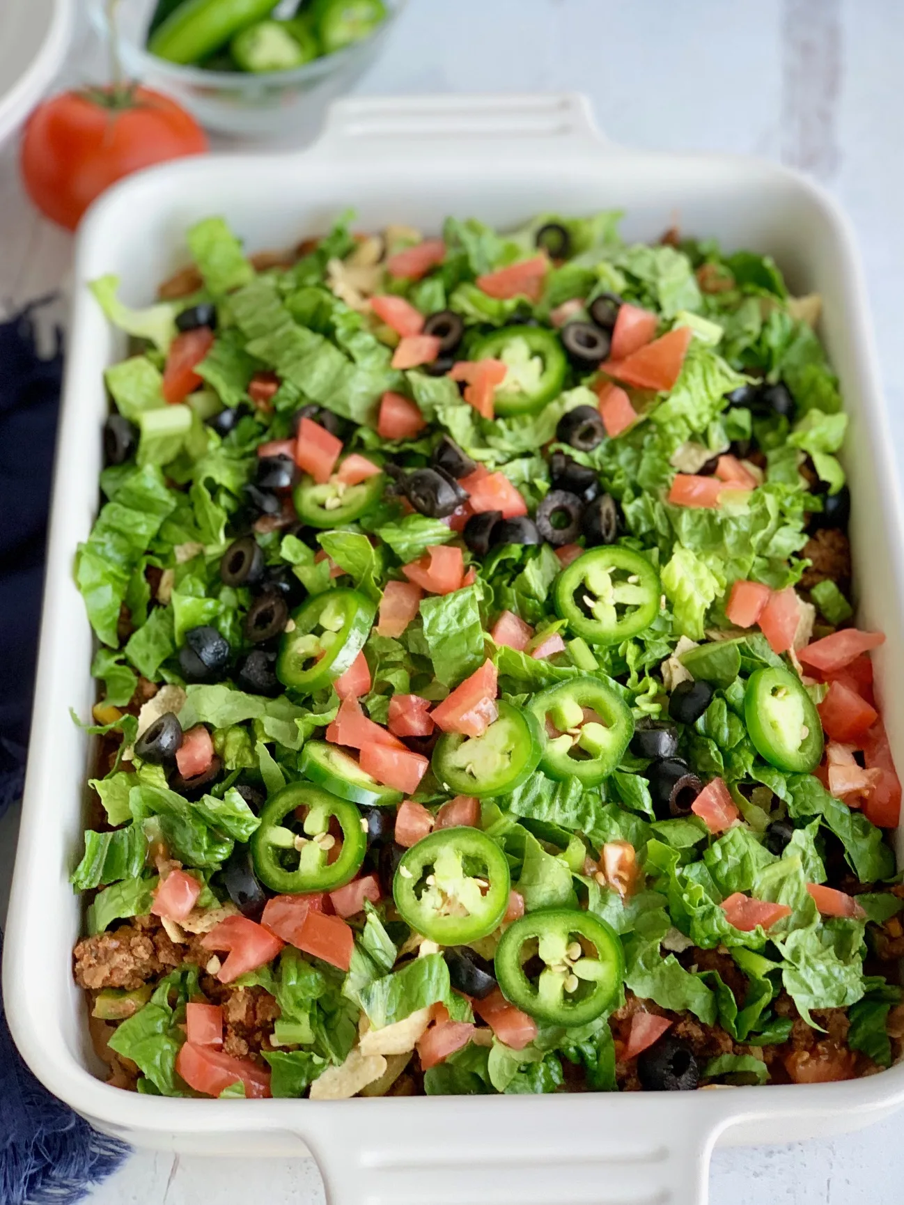 A baking dish filled with a layer of refried beans, taco meat sauce, crunched up chips, shredded romaine lettuce, diced tomatoes, jalapeno peppers, and black olives.