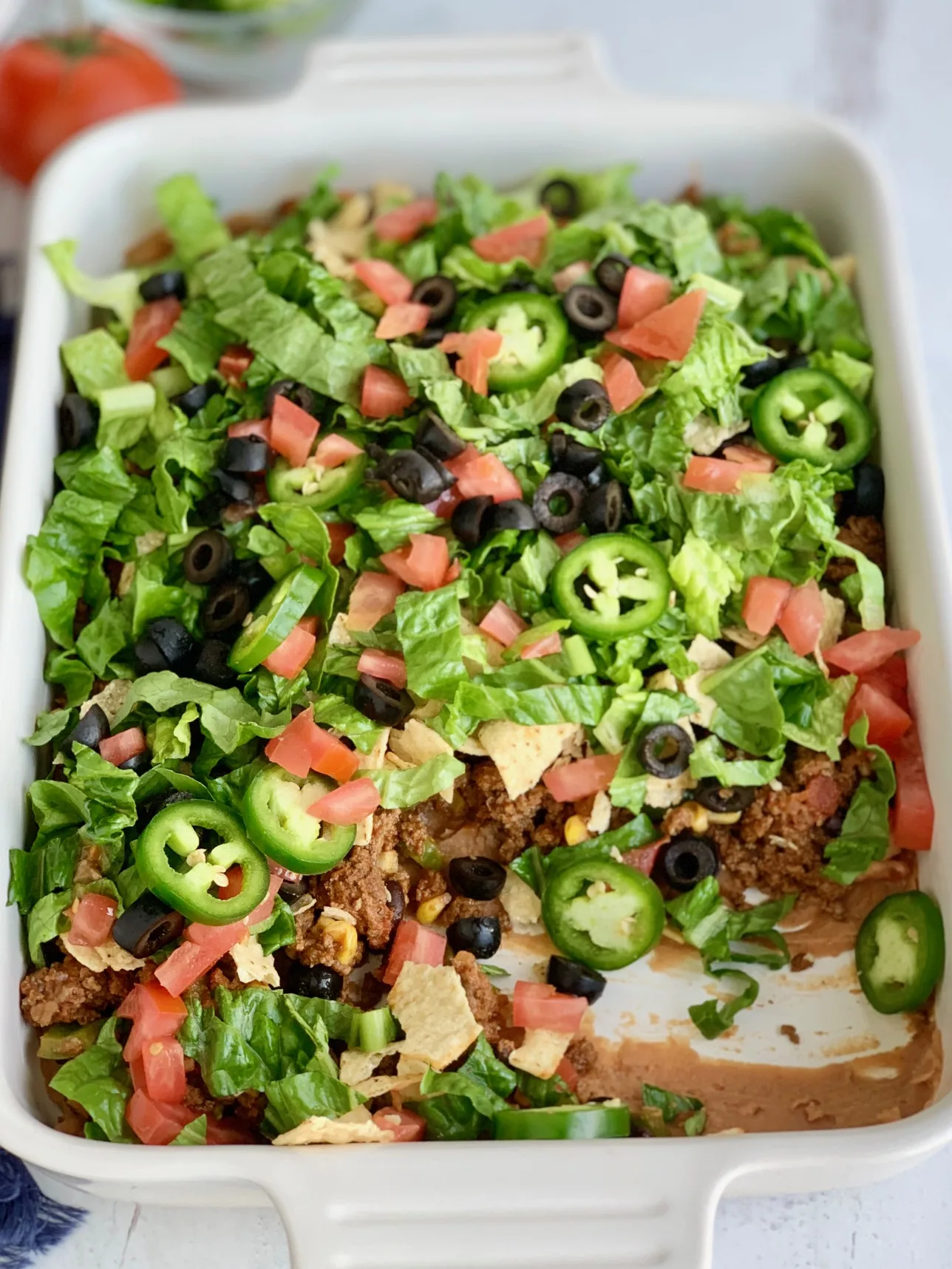 A baking dish filled with a layer of refried beans, taco meat sauce, crunched up chips, shredded romaine lettuce, diced tomatoes, jalapeno peppers, and black olives with some of it removed.