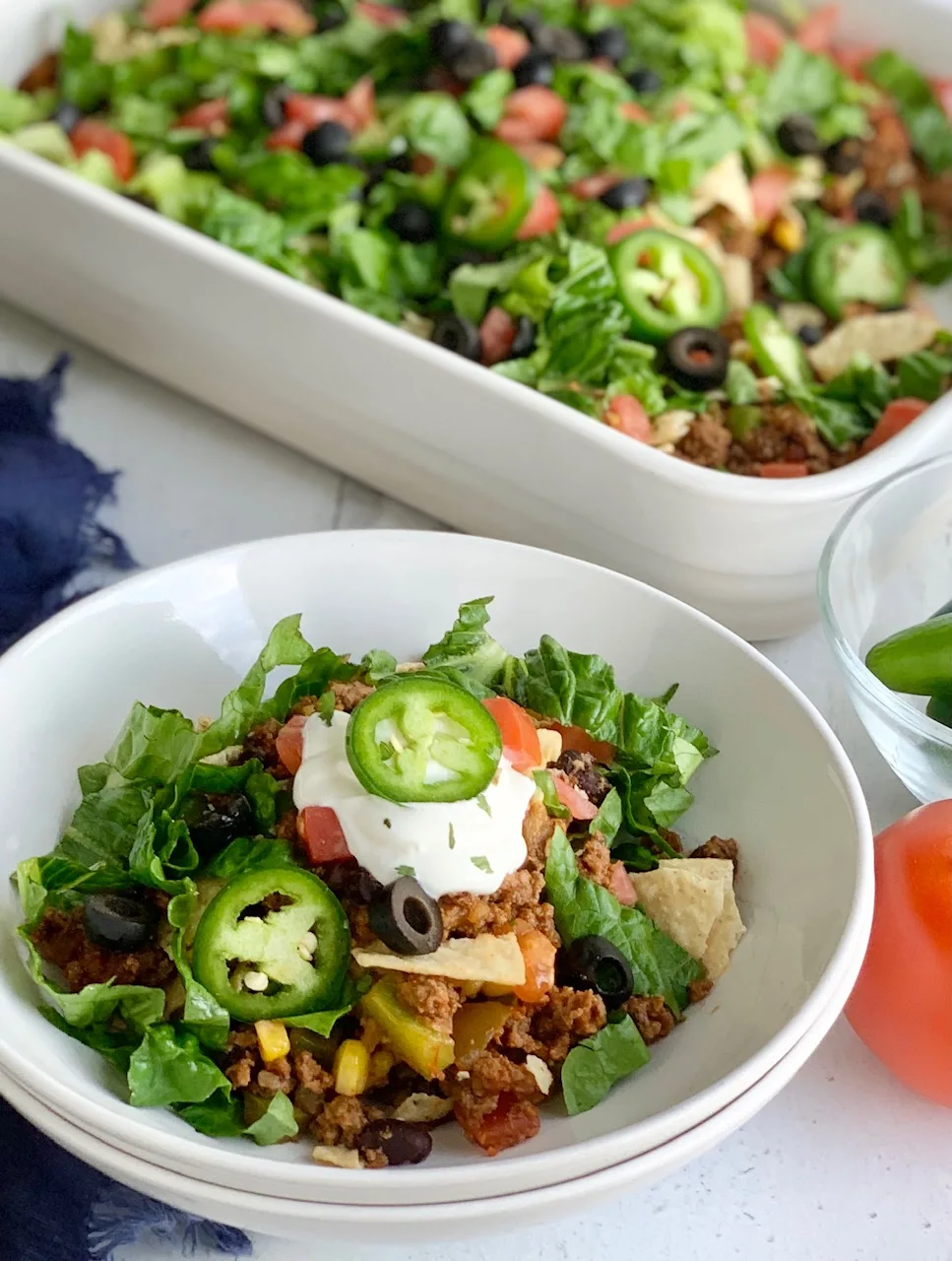A dinner bowl filled with refried beans, taco meat sauce, crunched up chips, shredded romaine lettuce, diced tomatoes, jalapeno peppers, and black olives. Then topped with a dollop of dairy free sour cream,