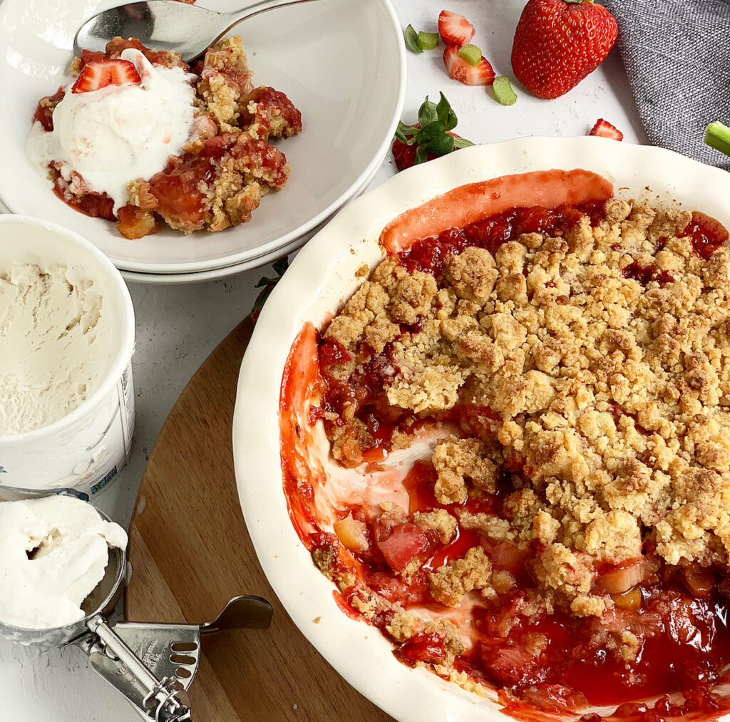 A large baking dish filled with baked strawberries and rhubarb with a crumble crust over the top next to a container of dairy free ice cream and serving bowls.