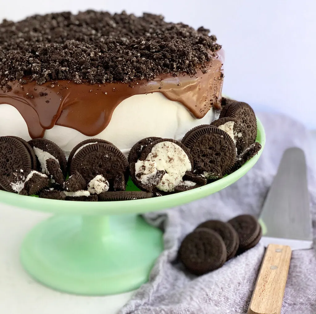 A round cake made from 2 layers of ice cream a surprise middle and covered in frozen cocowhip with homemade ganache running over the top and sides. Oreo like cookies are placed around the cake.