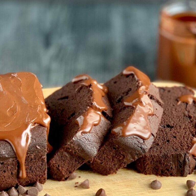 A loaf of chocolate loaf cake half cut and the slices laying on a cutting board. Chocolate ganache frosting is running down the top of each slice.