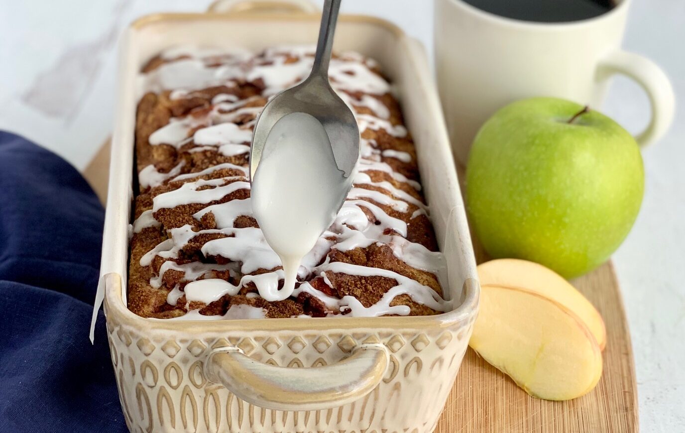 A loaf of bread still in the pan with a spoon drizzling white drizzle across the top in a zig zag motion next to a green Granny Smith apple and a cup of coffee.