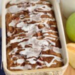 A loaf of bread still in the pan with a white drizzle zig zagging across the top of it next to a green Granny Smith apple and a cup of coffee.