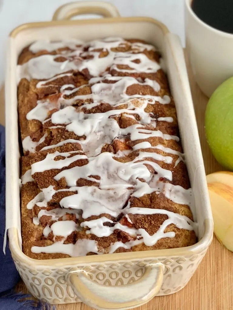 A loaf of bread still in the pan with a white drizzle zig zagging across the top of it next to a green Granny Smith apple and a cup of coffee.