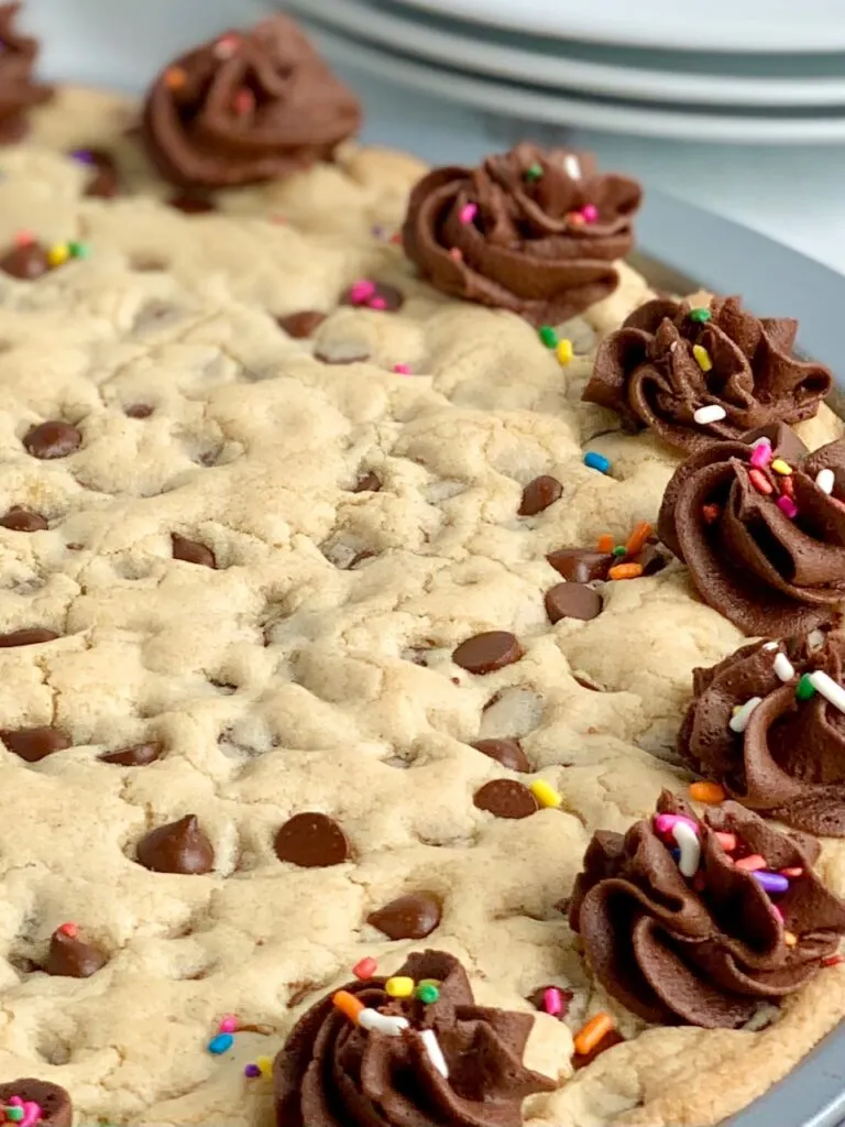 A close up of a giant chocolate chip cookie on a pizza pan with chocolate frosting swirls around the outside of the cookie. And colorful sprinkles are on top of the frosting. 