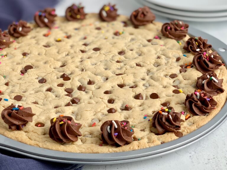 A giant chocolate chip cookie in a pizza pan with chocolate buttercream frosting swirls around the outside edge. And sprinkles are on top of it next to serving plates.
