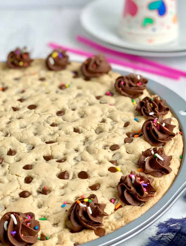 A giant chocolate chip cookie on a pizza pan with chocolate frosting swirls around the outside of the cookie. And colorful sprinkles are on top of the frosting. White plates with party cups on top of them are in the background.