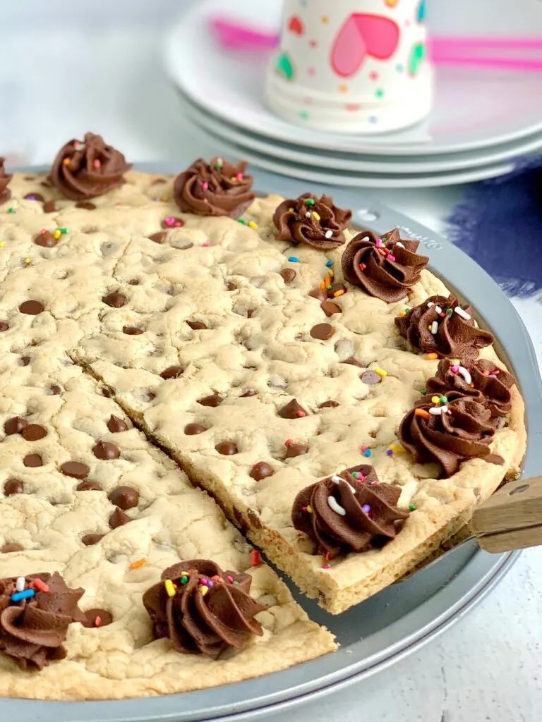 A giant chocolate chip cookie on a pizza pan with chocolate frosting swirls around the outside of the cookie. And colorful sprinkles are on top of the frosting. A slice has been cut and a pie server is removing it.