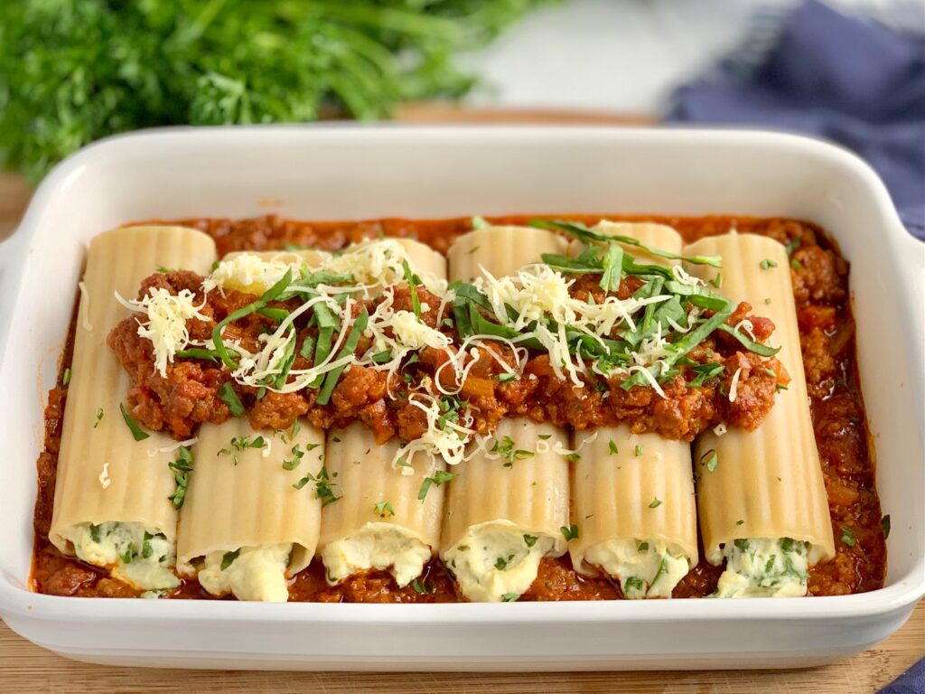 A white baking pan filled with ground beef and marinara topped with 6 soft round and long manicotti pasta shells. Each shell is filled with a creamy looking white ricotta mixture. Then more ground beef and marinara sauce is spread over that with dairy free shredded cheese and spinach on top of that.