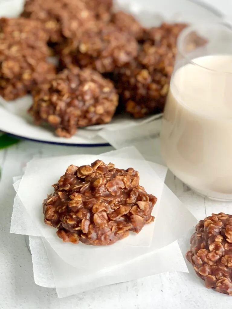 A close up of a No Bake Cookie with chocolate and peanut butter over oats next to a glass of milk, and another No Bake Cookie.