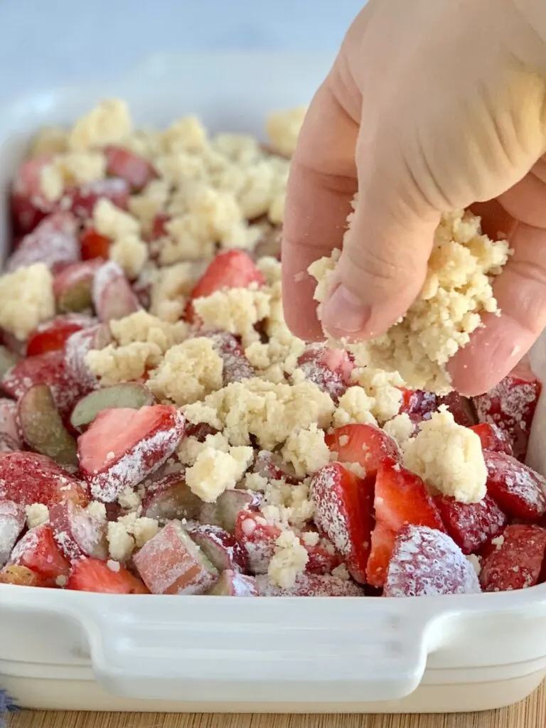 A white baking dish filled with diced fresh rhubarb and strawberries with a thick sweet juice and a hand is dropping a crumble topping on it.