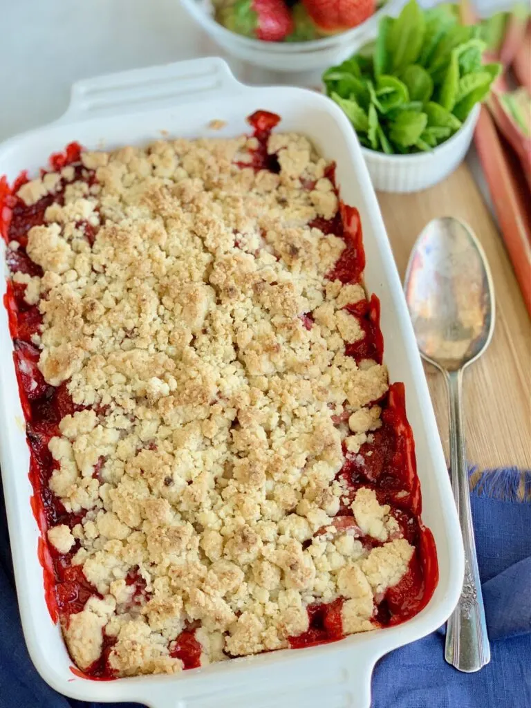 A white baking dish filled with diced fresh rhubarb and strawberries with a thick sweet juice and topped with a crumbly topping. There are two spoons in the dish and a scoop of vanilla ice cream with mint leaves.