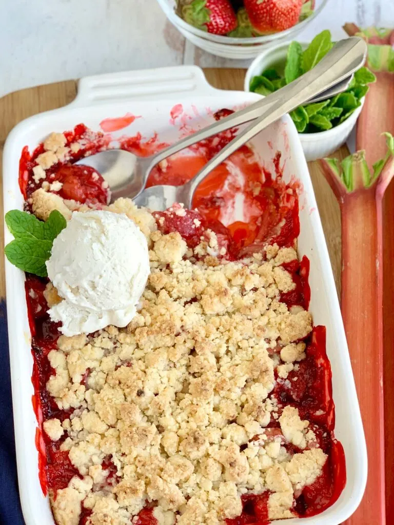 A white baking dish filled with diced fresh rhubarb and strawberries with a thick sweet juice and topped with a crumbly topping. There are two spoons in the dish and a scoop of vanilla ice cream with mint leaves.