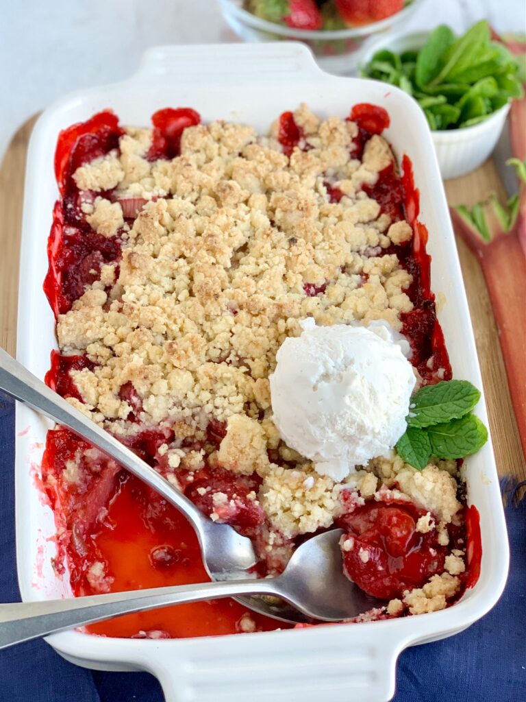 A white baking dish filled with diced fresh rhubarb and strawberries with a thick sweet juice and topped with a crumbly topping. There are two spoons in the dish and a scoop of vanilla ice cream with mint leaves.