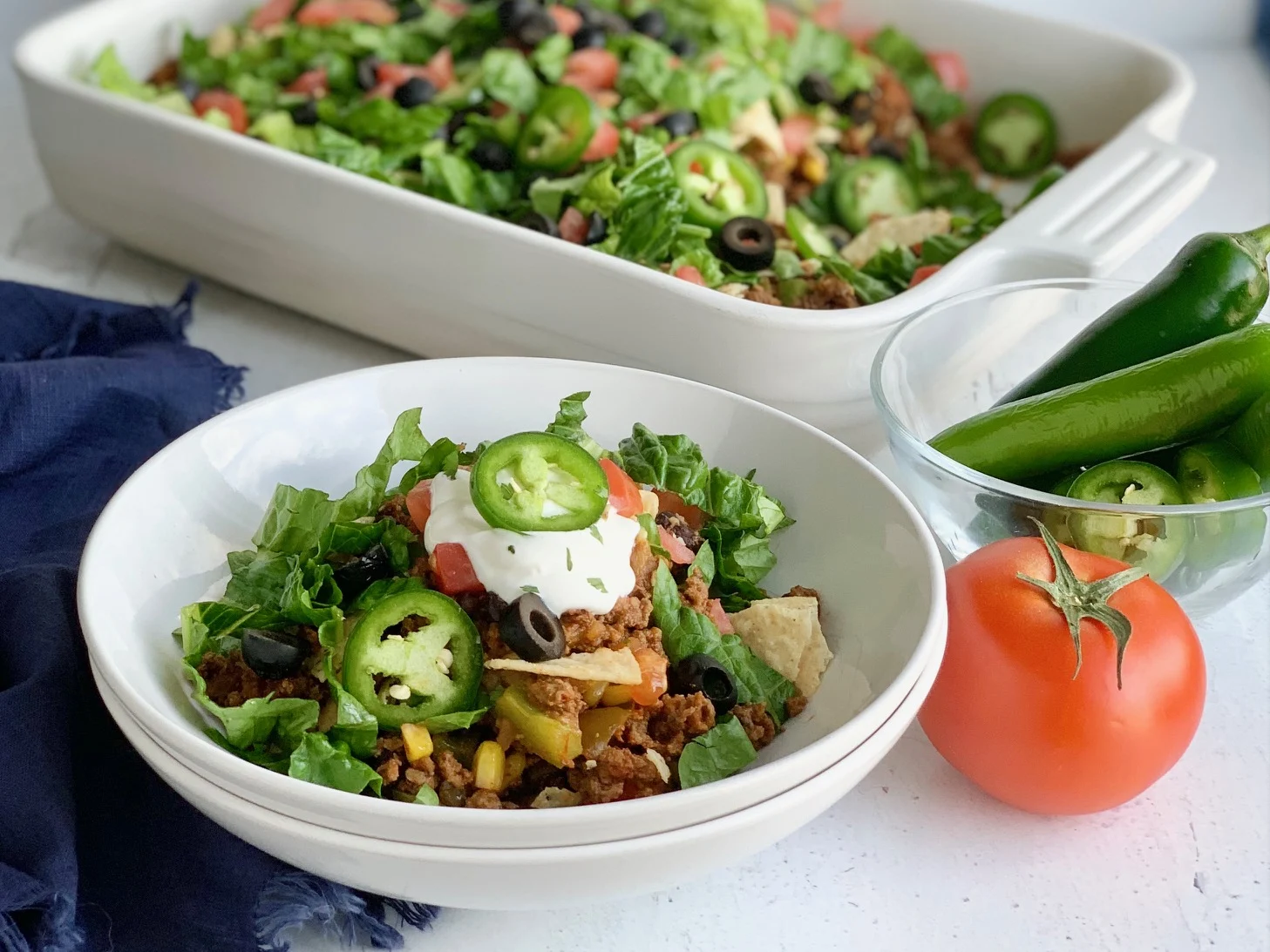 A white bowl holding a big helping of refried beans, seasoned ground beef, diced tomatoes and olives topped with dairy free sour cream and jalapenos.