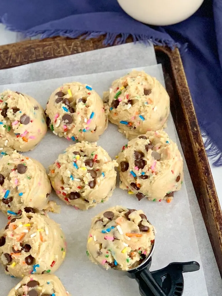 A baking sheet full of round raw dough balls of chocolate chip cookie dough with colorful sprinkles. A cookie scooper is full of dough ready to place onto the cookie sheet.