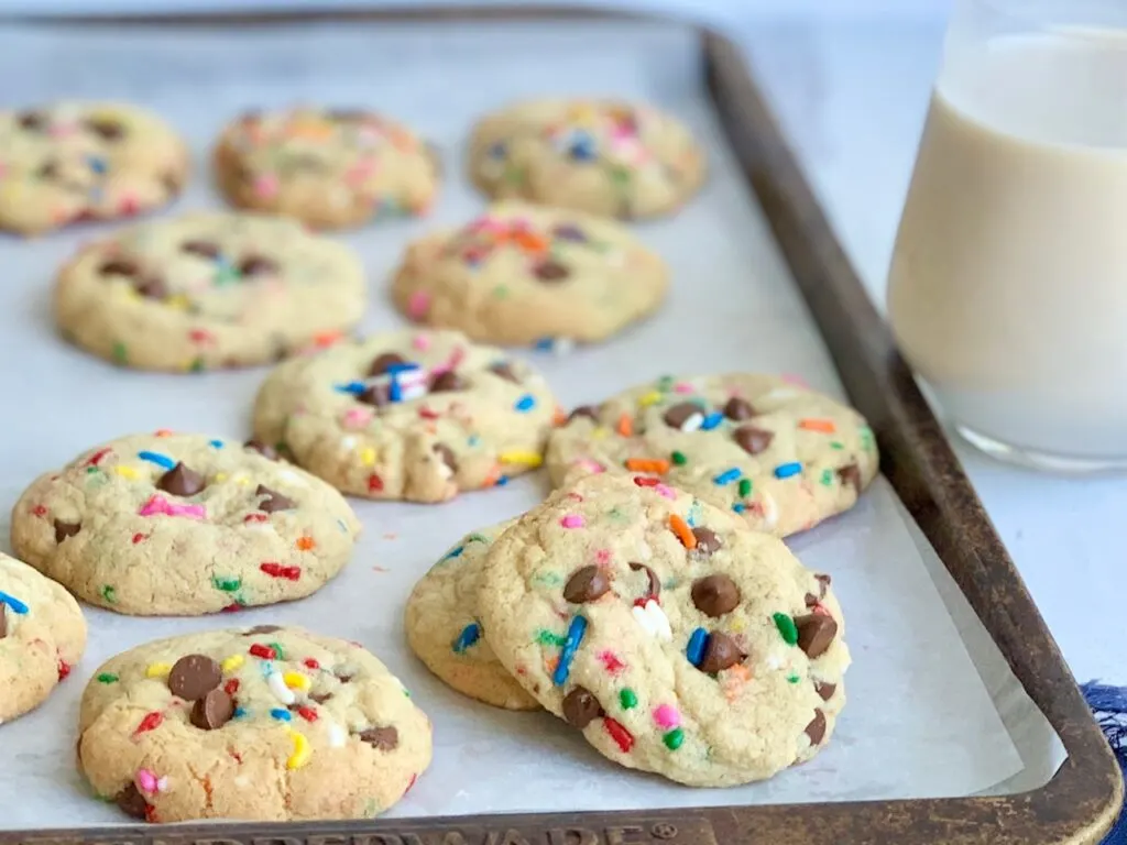 A baking sheet filled with chocolate chip cookies with colorful sprinkles next to a glass of dairy free milk.