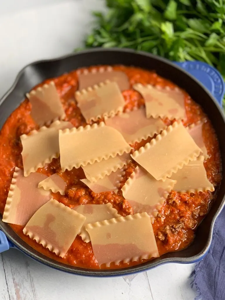 A skillet filled with broken lasagna noodles in a meaty marinara sauce next to a bunch of fresh parsley.