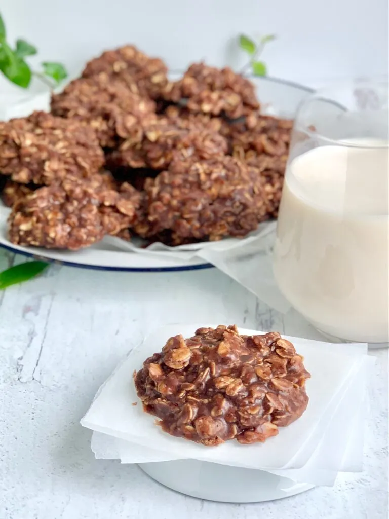 A close up of a No Bake Cookie with chocolate and peanut butter over oats next to a glass of milk. In the background is a cookie plate full of No Bake Cookies.