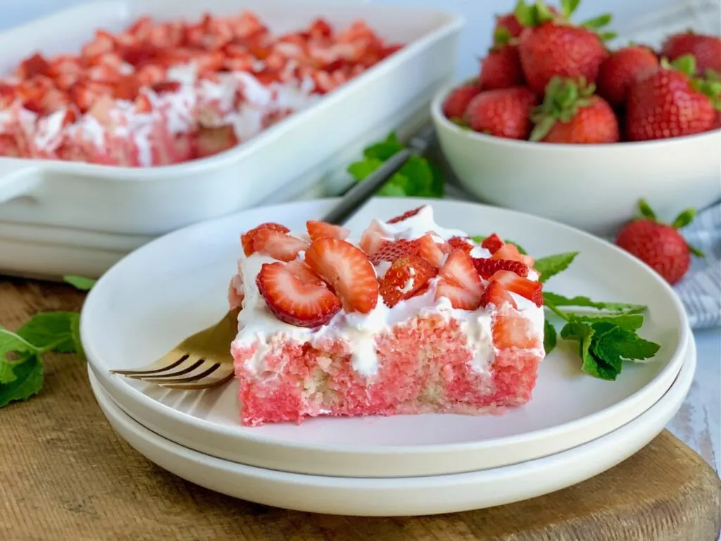 A dessert plate with a large slice of cake with jello in it with a whipped topping layer and fresh cut strawberries next to a fork on a cutting board.