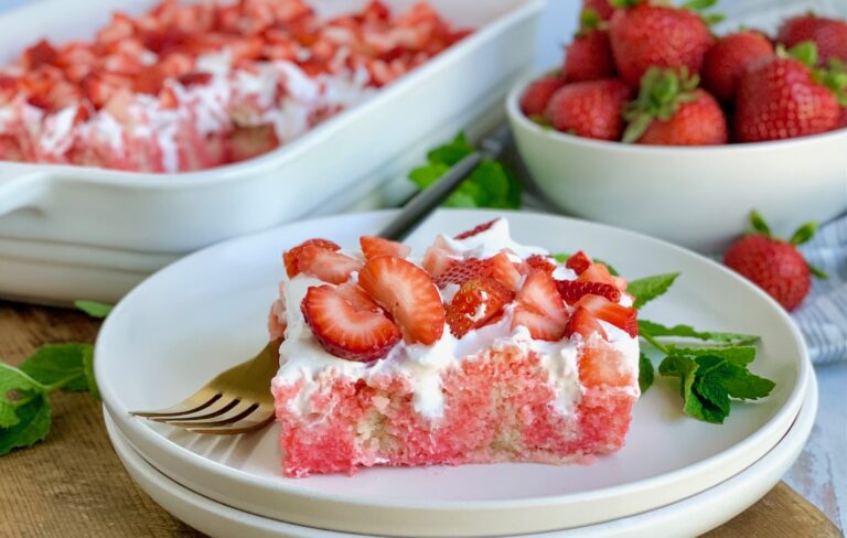 A large slice of vanilla cake with strawberry jello in it with a whipped topping on it and fresh strawberries on that. There is a fork nearby and a bowl of strawberries.