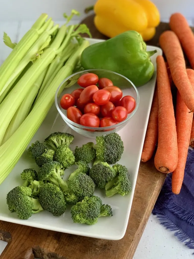 A large white plate filled with a bunch of celery, a green and yellow bell pepper, large raw carrots, a bowl of cherry tomatoes, and pieces of broccoli.