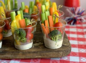 A small party drink cup with white ranch dressing in the bottom. Then sliced celery, carrots, bell peppers in the back of the cup. 3 cherry tomatoes on a fun bamboo skewer in front with a piece of broccoli.