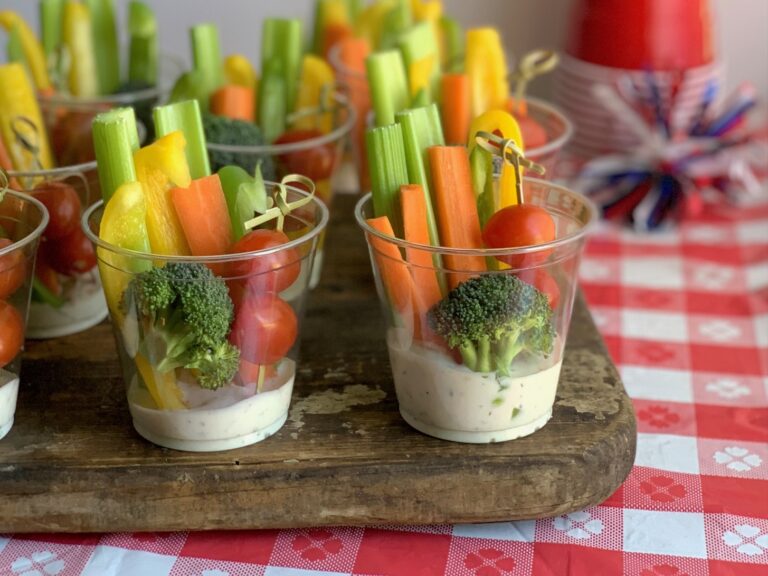 A small party drink cup with white ranch dressing in the bottom. Then sliced celery, carrots, bell peppers in the back of the cup. 3 cherry tomatoes on a fun bamboo skewer in front with a piece of broccoli.