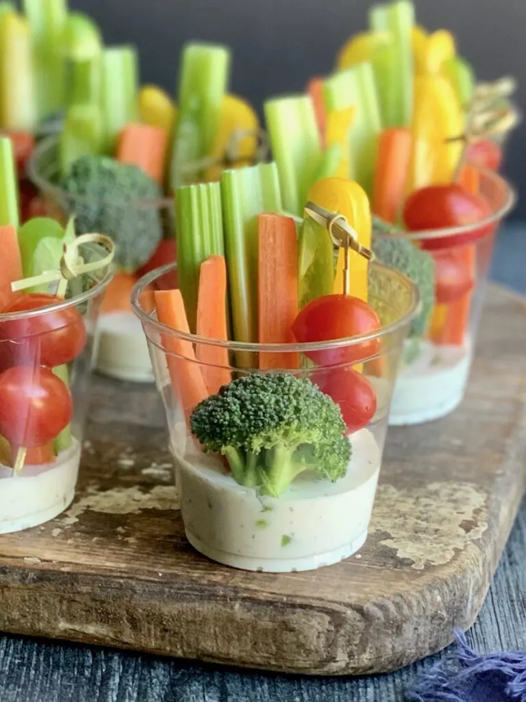 Small party drink cups with white ranch dressing on the bottom of the cup. Celery, carrots, and yellow bell pepper slices are in the back of the cups. Then 3 cherry tomatoes are  on a bamboo skewer next to a piece of broccoli.