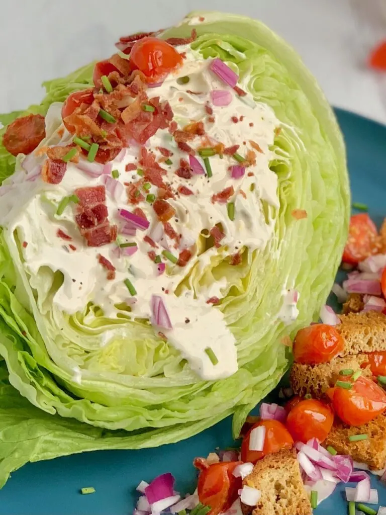 A quartered slice of iceberg lettuce with ranch dressing drizzled over. Then topped with bacon crumbles, cherry tomatoes cut in half, croutons, red onion slices, and small bits of green onions on a blue salad plate.