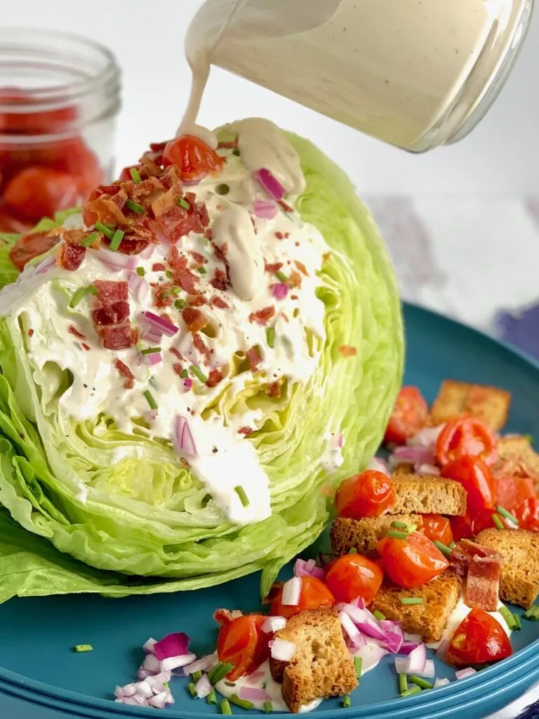 A quartered slice of iceberg lettuce with someone drizzling ranch dressing drizzled on top of the salad. Then topped with bacon crumbles, cherry tomatoes cut in half, croutons, red onion slices, and small bits of green onions on a blue salad plate.