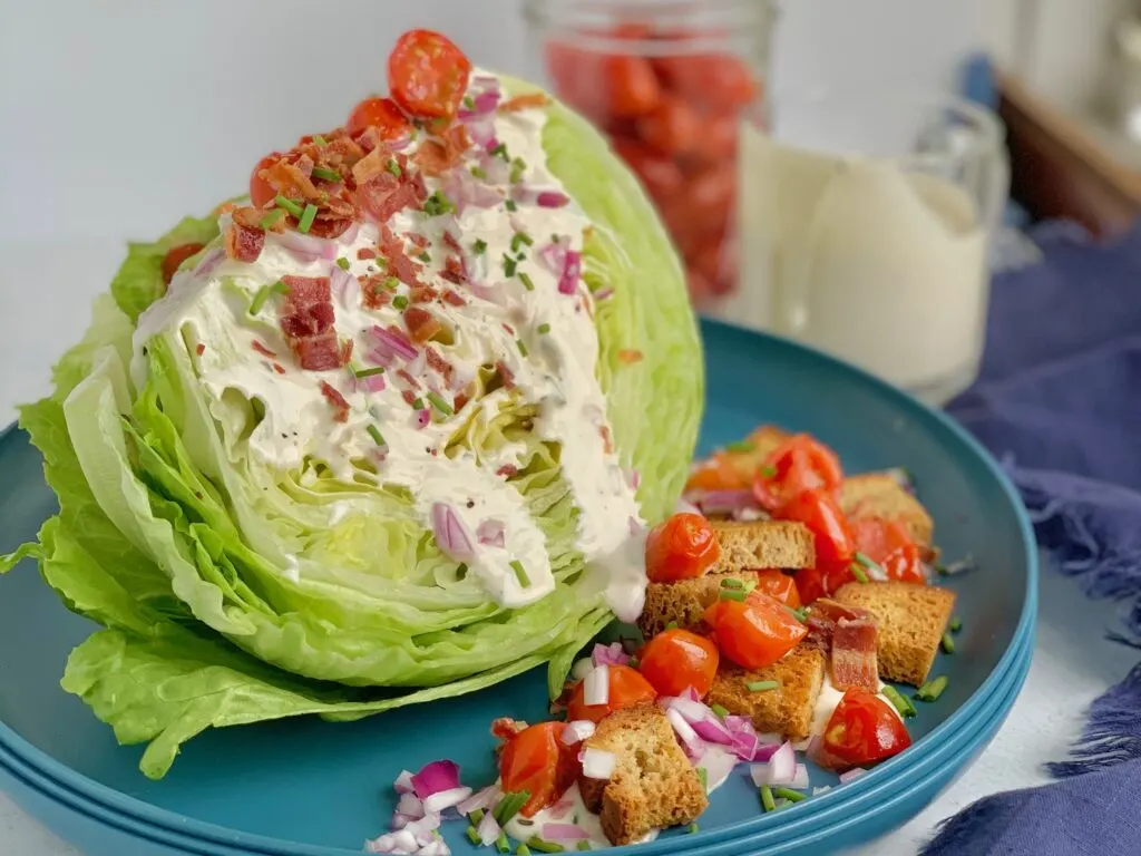 A quartered slice of iceberg lettuce with ranch dressing drizzled over. Then topped with bacon crumbles, cherry tomatoes cut in half, croutons, red onion slices, and small bits of green onions on a blue salad plate.