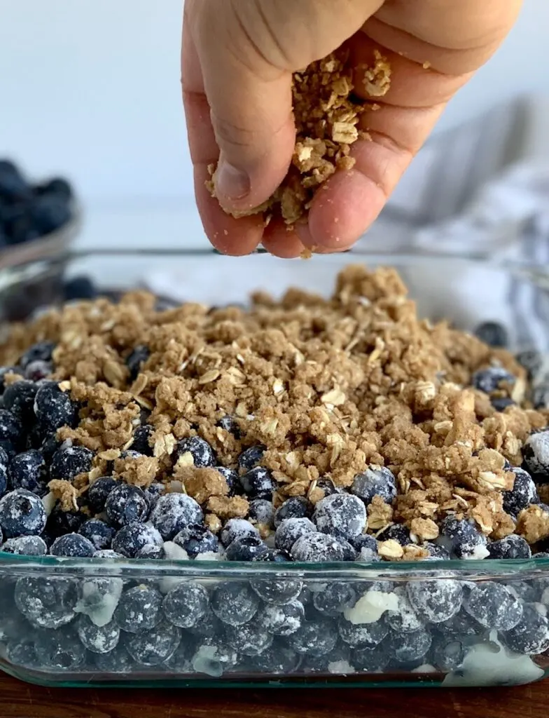 A baking dish filled with blueberries coated in corn starch. And a hand is dropping the crisp topping over the blueberries.