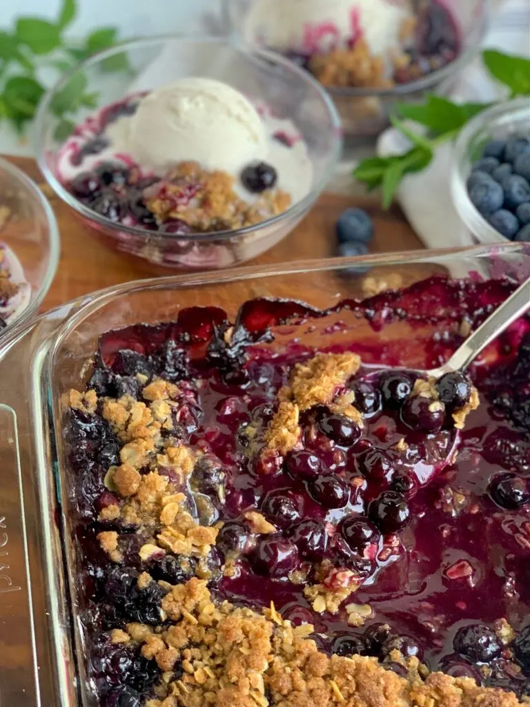 A glass baking dish filled with a thick blueberry mixture on the bottom and a golden oat, flour, butter mixture on top. There are also bowl in the background filled with the mixture and ice cream.