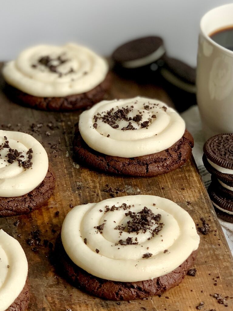 A brown brownie cookie with vanilla buttercream on top in s swirl design with crushed oreo cookie bits on top on a dark cutting board next to a mug of coffee. 