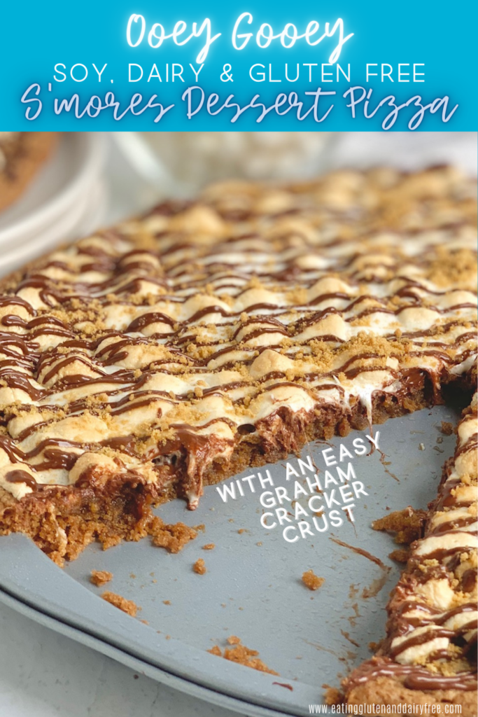 A pizza pan layered with a graham cracker crust, melted chocolate, golden brown mini marshmallows, then drizzled with chocolate and topped with graham cracker crumbs. A large slice is removed from the pizza.