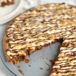 A pizza pan layered with a graham cracker crust, melted chocolate, golden brown mini marshmallows, then drizzled with chocolate and topped with graham cracker crumbs. A large slice is removed from the pizza and placed on a plate nearby.