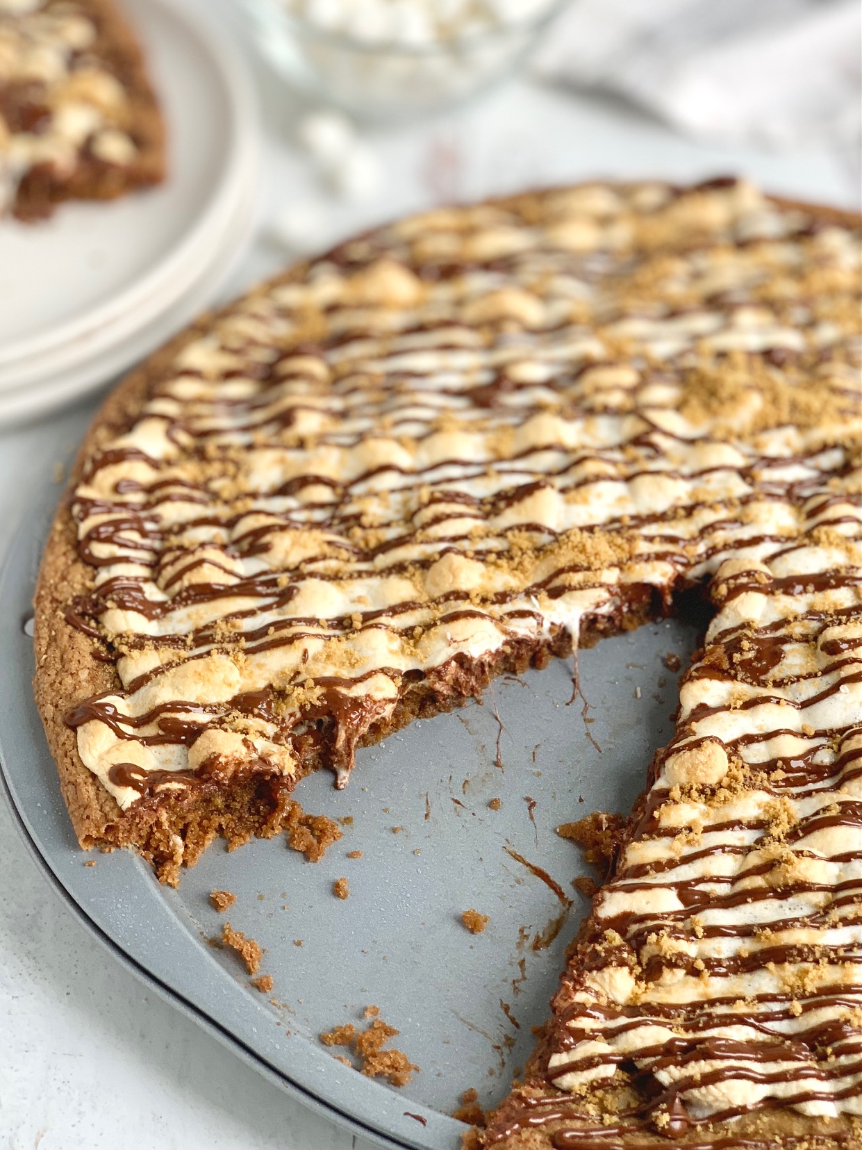 A pizza pan layered with a graham cracker crust, melted chocolate, golden brown mini marshmallows, then drizzled with chocolate and topped with graham cracker crumbs. A large slice is removed from the pizza and placed on a plate nearby.