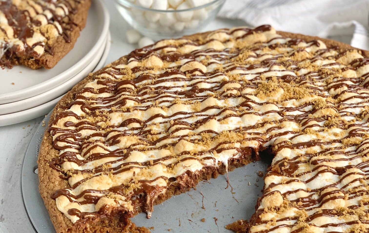 A pizza pan filled with a chewy graham cracker crust, melted chocolate chips, ooey gooey golden brown mini marshmallows, melted chocolate drizzled across the top, and last topped with a handful of crushed graham crackers. This is next to a set of dessert plates and a bowl full of mini marshmallows.