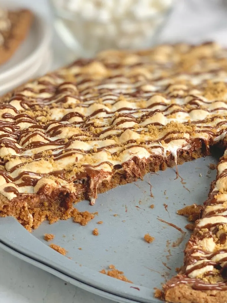 A pizza pan layered with a graham cracker crust, melted chocolate, golden brown mini marshmallows, then drizzled with chocolate and topped with graham cracker crumbs. A large slice is removed from the pizza and placed on a plate nearby.