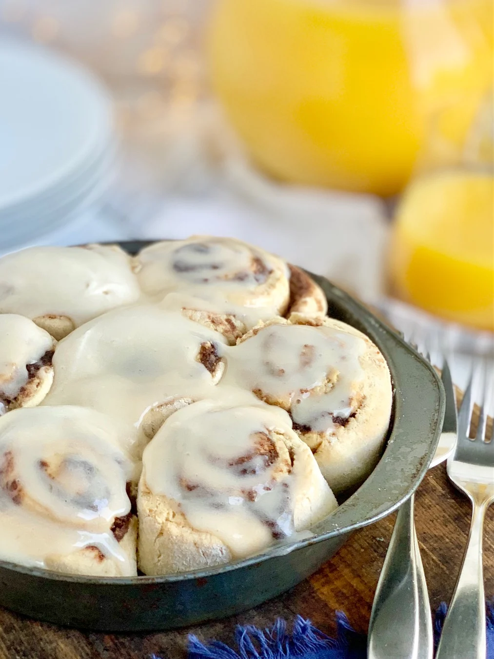 A pie plate filled with soft, gooey cinnamon rolls and topped with a decadent creamy dairy free cream cheese frosting next to serving plates and glass of orange juice.