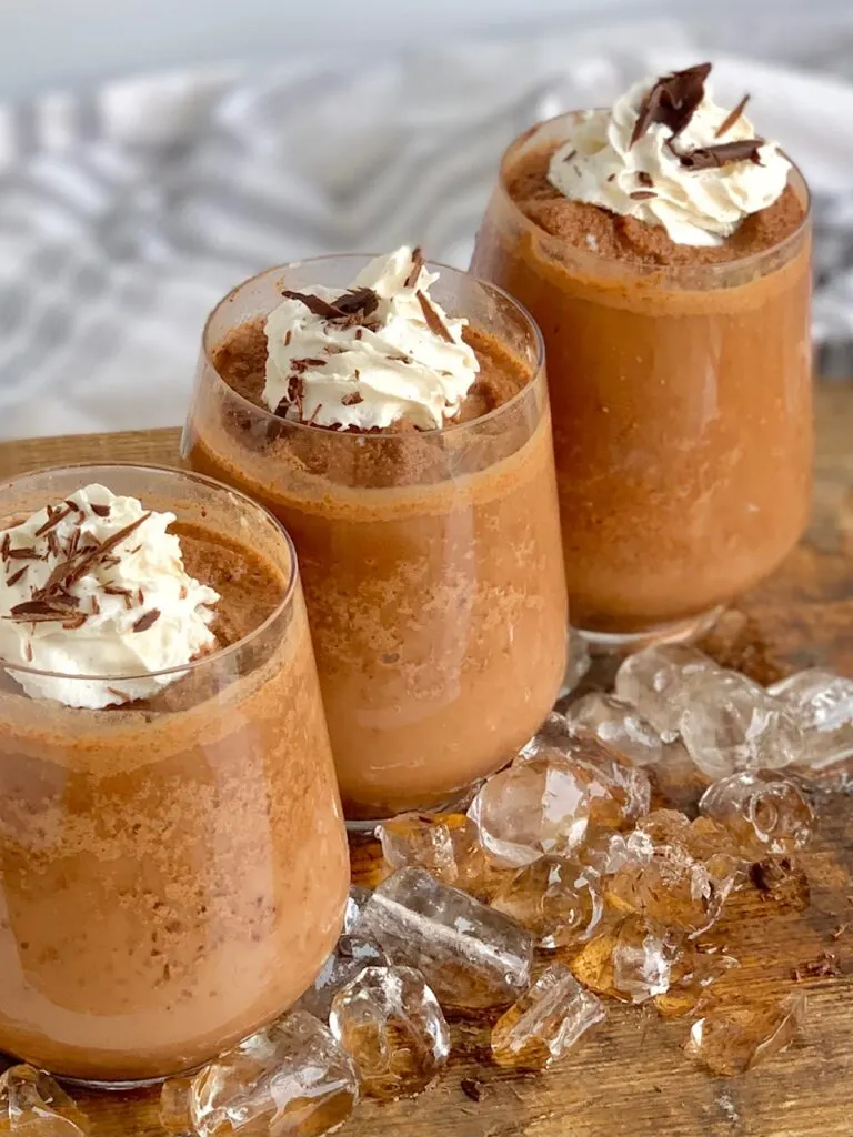Three decorative glasses filled with a chocolatey cocoa mixture topped with whipped cream and chocolate shavings next to cubed ice.