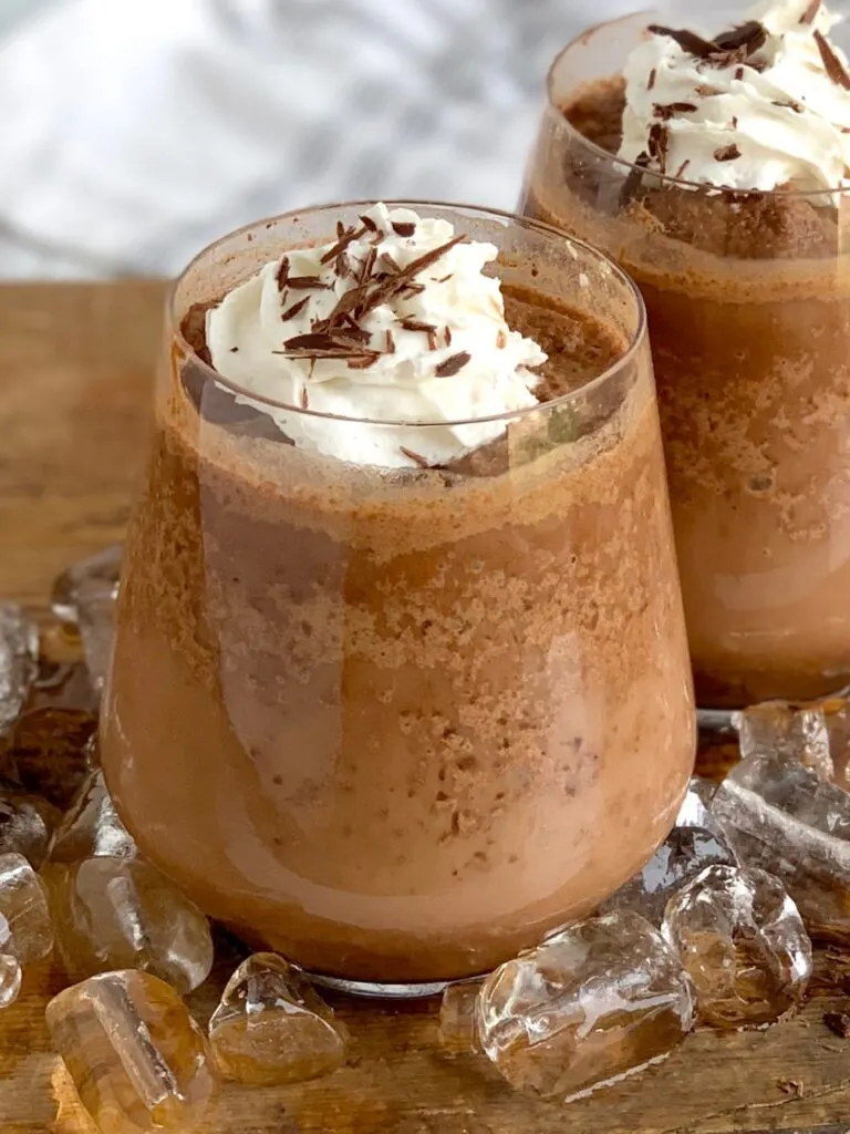 A close up of a decorative glass filled with a chocolatey cocoa mixture topped with whipped cream and chocolate shavings next to cubed ice.