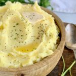 A large wooden bowl with mashed potatoes topped with melting butter and peper.