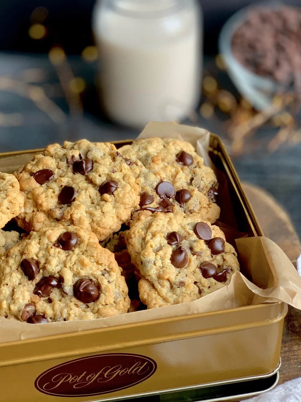 A gold colored cookie tin packed full of golden brown cookies loaded with chocolate chips next to a cup of milk.