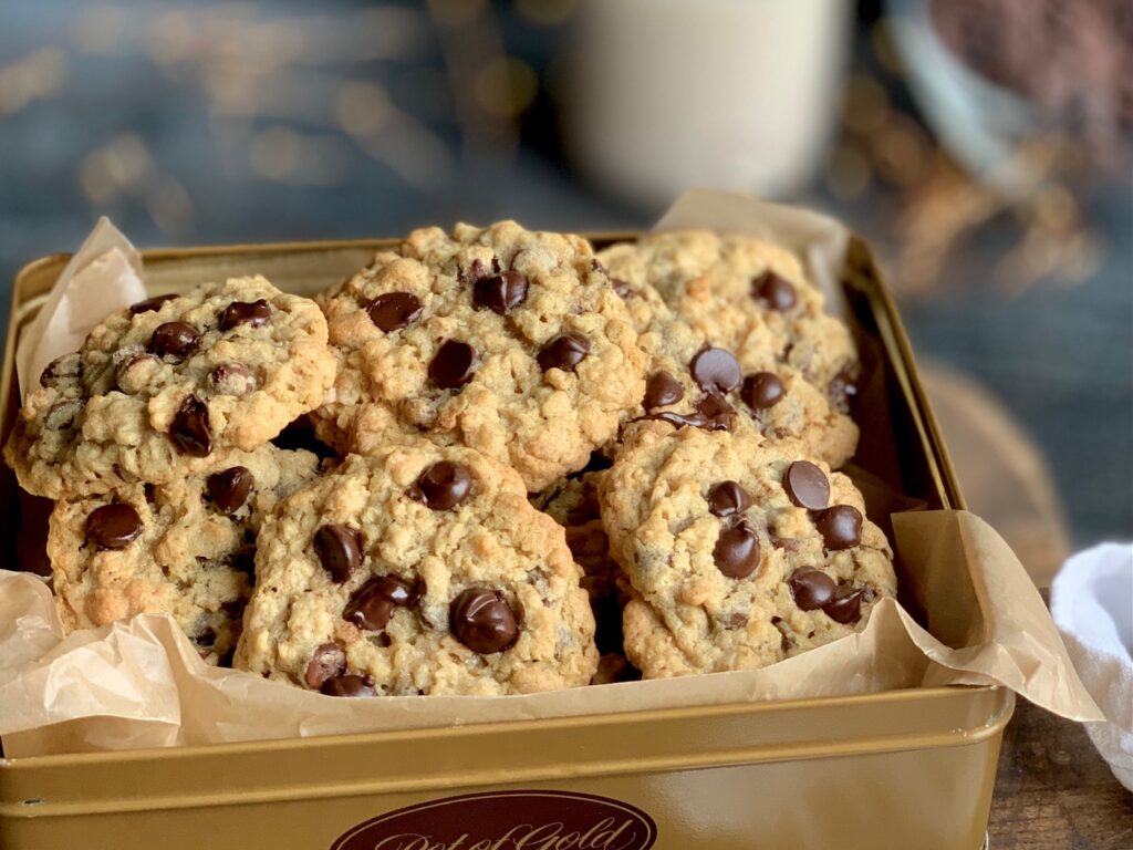 A gold colored cookie tin packed full of golden brown cookies loaded with chocolate chips next to a cup of milk.