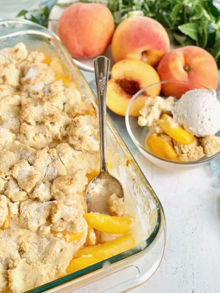 A 9x13-inch baking dish filled with a thick sweet peach mixture and topped with a biscuit-like golden brown dough. With sprinkles of sugar and a dessert nearby with cobbler in it and a scoop of ice cream.