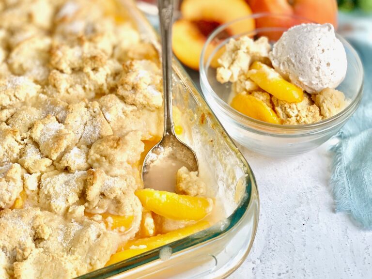 A baking dish filled with sweetened peached on the bottom and layered with biscuit style topping next to a dessert bowl filled with cobbler and a scoop of ice cream.